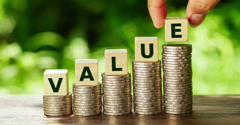 Five stacks of coins rise from left to right with wooden blocks spelling "VALUE" placed on top. A hand adds the final block to the tallest stack to represent the growing value of personal branding over time.