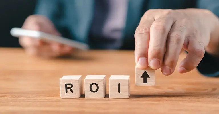 A business professional places a wooden block with an upward arrow beside blocks spelling "ROI" while holding a smartphone in the background. The scene represents the value of personal branding through increased visibility and measurable return on investment.