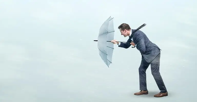 A business professional leans forward while gripping an inside out umbrella in strong wind against a pale background. The struggle represents the value of personal branding when building resilience and maintaining a clear professional identity during challenges.