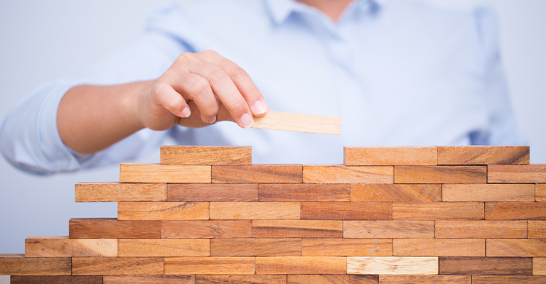 A person places a wooden block onto a growing wall of stacked blocks, symbolizing building experience and developing professional skills. The image represents the step by step process of preparing for success with LinkedIn for internships and career growth.